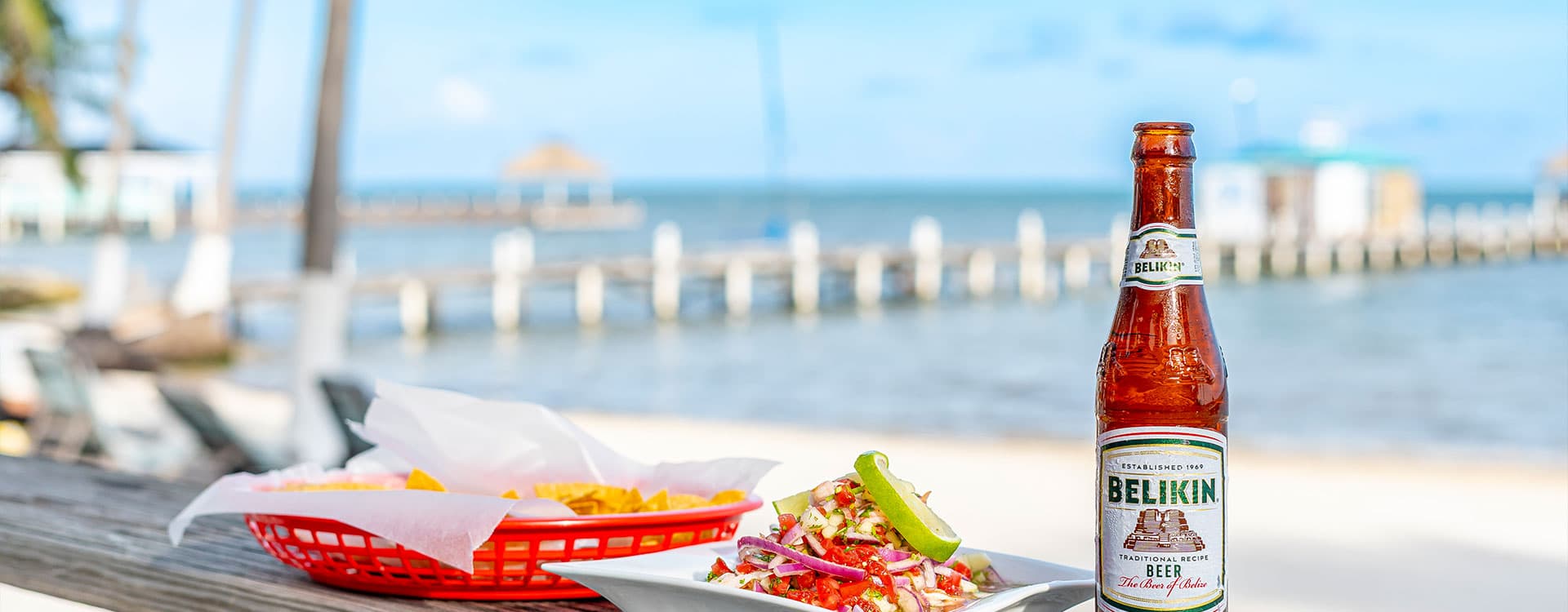A bottle of Belikin beer beside a plate of nachos and a colorful salad, set against a beach view with a dock.
