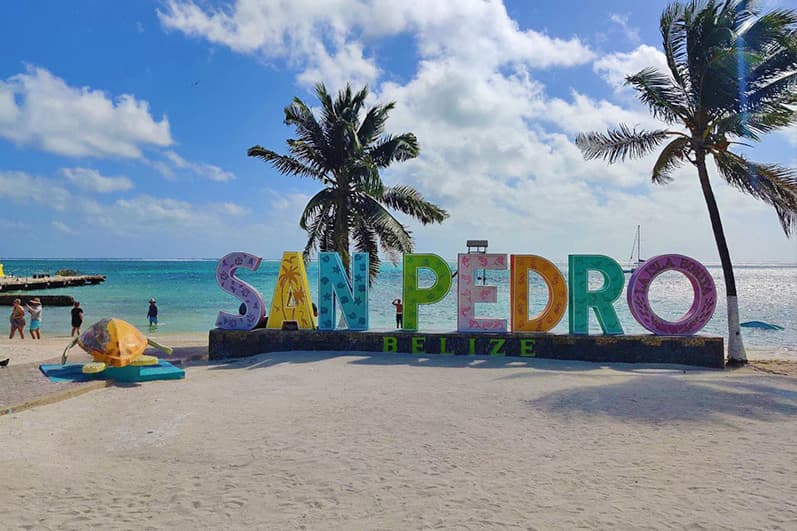 Colorful "San Pedro, Belize" sign on a sandy beach with palm trees and a clear blue sky.